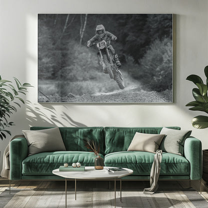 A dynamic black and white photograph of a motocross racer in mid-air, jumping over a dirt mound. The rider, wearing full protective gear and a helmet, is on a dirt bike with the number 19 plate. The background is a soft-focus forest, and the entire image is enclosed in a silver frame. Poster