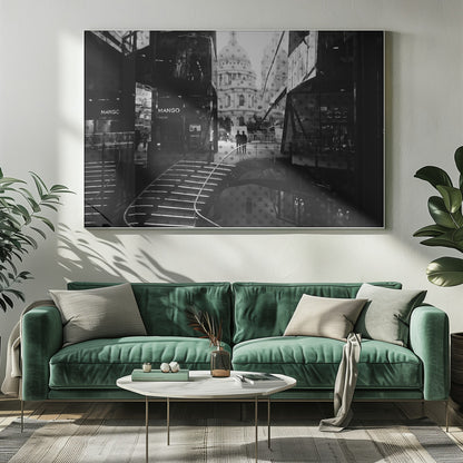 A framed, black and white photograph capturing a layered urban scene. The view, seen through a window with reflections of a curved, lit staircase, shows two people walking towards a large domed cathedral framed by modern glass buildings. Wall Art