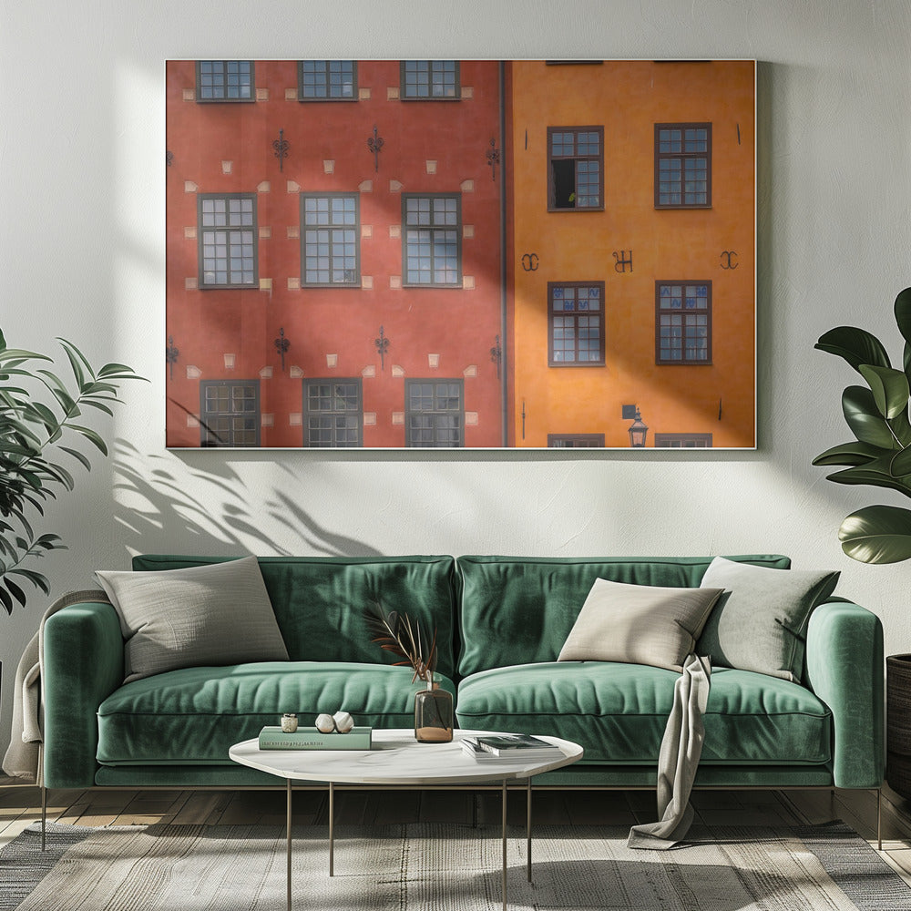 A framed close-up photograph of two adjacent, colorful building facades. The building on the left is a deep, rustic red, while the one on the right is a vibrant orange. Both feature numerous multi-paned windows with dark frames. Artwork