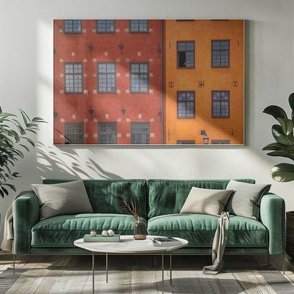 A framed close-up photograph of two adjacent, colorful building facades. The building on the left is a deep, rustic red, while the one on the right is a vibrant orange. Both feature numerous multi-paned windows with dark frames. Artwork