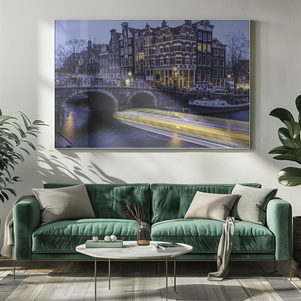 A long-exposure photograph of an Amsterdam canal at dusk, featuring a bright yellow light trail from a passing boat. In the background, illuminated historic canal houses line the water, and an arched stone bridge covered in bicycles crosses the canal. The entire image is presented in a silver frame. Print