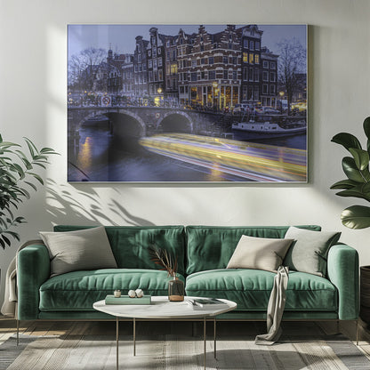A long-exposure photograph of an Amsterdam canal at dusk, featuring a bright yellow light trail from a passing boat. In the background, illuminated historic canal houses line the water, and an arched stone bridge covered in bicycles crosses the canal. The entire image is presented in a silver frame. Print