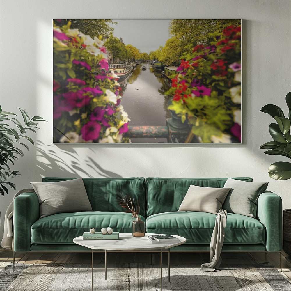 A scenic view looking down a canal in Amsterdam from a bridge, framed by vibrant, out-of-focus purple, red, and white flowers in the foreground. The canal is lined with houseboats and lush green and yellow trees, with historic buildings visible behind them. Poster
