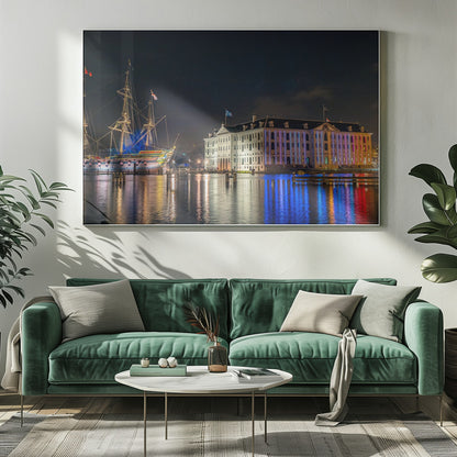 A vibrant nighttime photograph of the National Maritime Museum and a replica of the East Indiaman Amsterdam ship in a harbor. Both are illuminated with colorful lights that cast beautiful reflections on the dark water. Wall Art