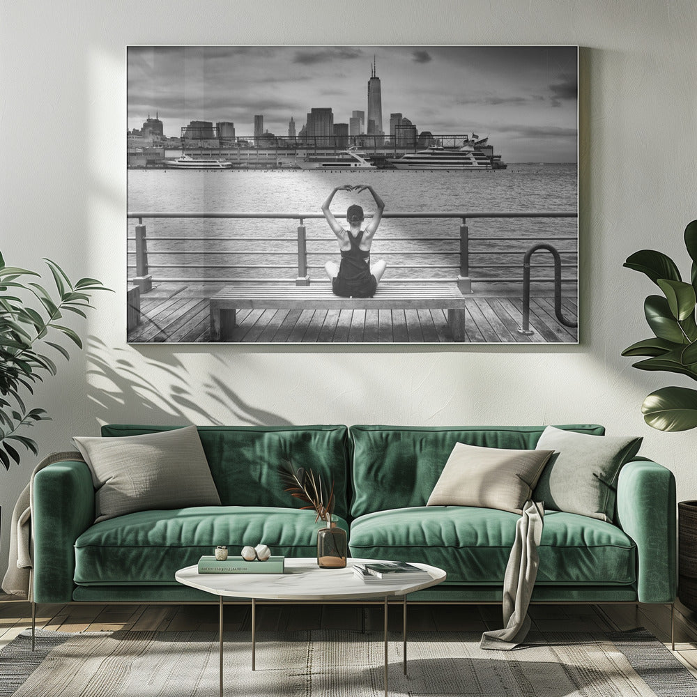 A framed black and white photograph showing the back of a woman sitting on a wooden bench on a pier. She is in a yoga pose with her arms raised, looking out at the Lower Manhattan skyline, including the Freedom Tower, across the water. Decor
