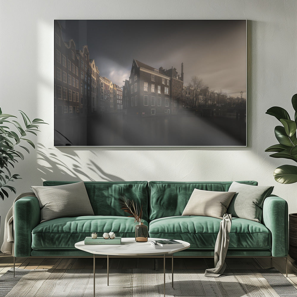 A moody, atmospheric photograph of historic canal houses in Amsterdam, viewed from the water. The scene is dark, with a dramatic, cloudy sky and a soft glow on the horizon, while some windows in the dark brick buildings are lit from within. The image is bordered by a silver frame. Wall Art