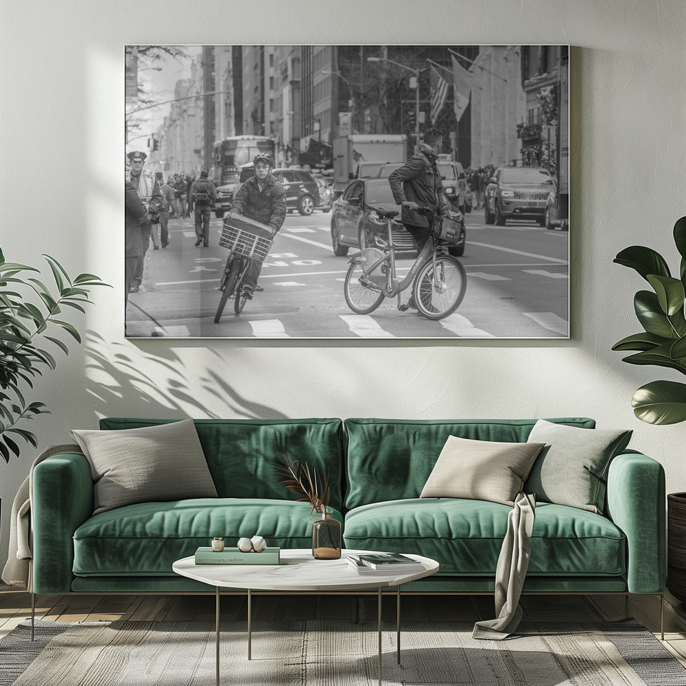 A black and white street photograph of a busy city intersection, framed in silver. In the foreground, two men are crossing with their bicycles amidst traffic, with one cyclist looking directly at the camera. Tall buildings and other pedestrians line the background, capturing a moment of everyday city life. Artwork