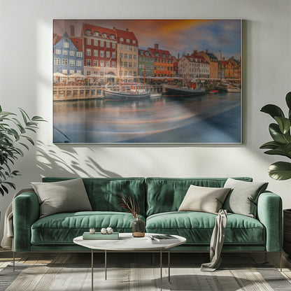 A long-exposure photograph of the colorful Nyhavn harbor in Copenhagen at sunset. Historic, brightly colored buildings line the canal, where two traditional wooden boats are moored. The sky is a dramatic blend of orange and blue, and the water is blurred, showing streaks of reflected light. Decor