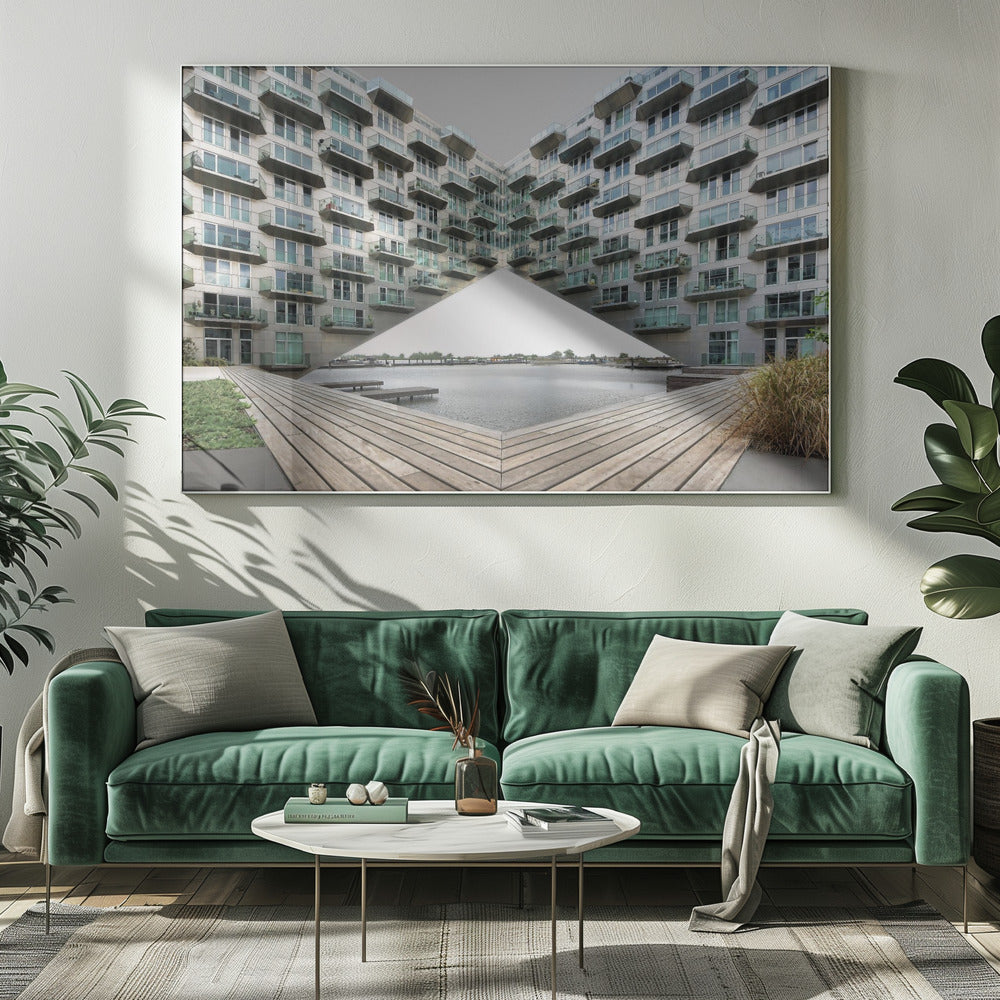 A symmetrical, mirrored photograph of a modern V-shaped apartment complex with numerous glass balconies, overlooking a wooden deck and a calm body of water under a grey sky. The image is presented within a silver picture frame. Artwork