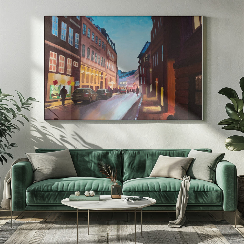 An impressionistic painting of a European city street at dusk, shown in a silver frame. The sky is a bright, clear blue, contrasting with the deep brown and maroon buildings. Warm yellow light glows from shop windows and street lamps, reflecting on the wet-looking street where cars are parked. Decor