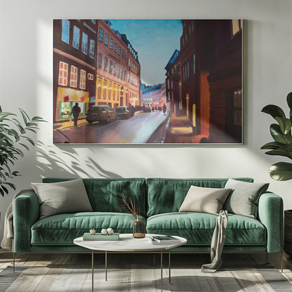 An impressionistic painting of a European city street at dusk, shown in a silver frame. The sky is a bright, clear blue, contrasting with the deep brown and maroon buildings. Warm yellow light glows from shop windows and street lamps, reflecting on the wet-looking street where cars are parked. Decor