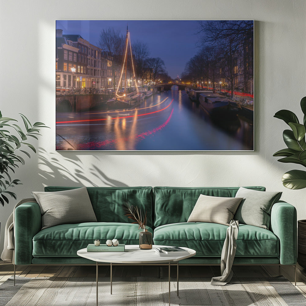 A long-exposure photograph of an Amsterdam canal at dusk. The scene is bathed in a deep blue light, with warm yellow and orange lights from street lamps and buildings reflecting on the water. A sailboat is decorated with glowing string lights, and red and white light trails from a passing boat create streaks across the canal. Houseboats are moored along the sides, and a bridge is visible in the distance. Poster