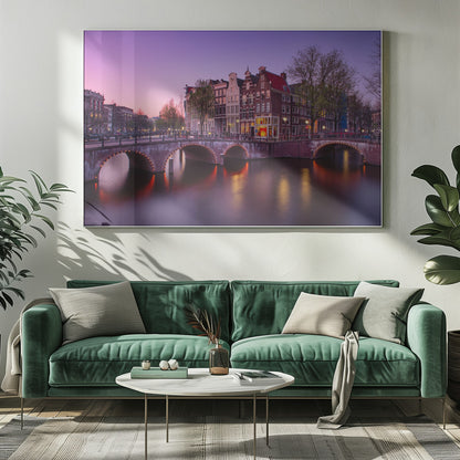 A scenic view of an Amsterdam canal at twilight, with historic brick buildings and illuminated arched bridges reflected in the calm water under a vibrant pink and purple sky. Poster