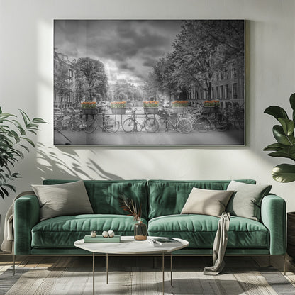 A selective color, black and white photograph of a classic Amsterdam canal scene, enclosed in a silver frame. The foreground shows a bridge lined with numerous bicycles and planters filled with vibrant orange tulips, which are the only colored elements. The background features canal houses, trees, and another bridge under a dramatic, cloudy sky with sunbeams. Print