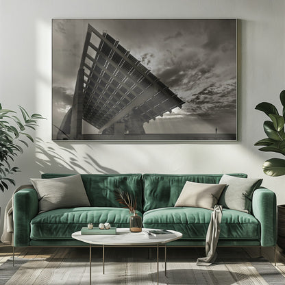 A framed, dramatic black and white photograph showing a massive, tilted solar panel structure from a low angle. The sky is filled with dark, moody clouds with some sun rays breaking through. A tiny silhouette of a person in the lower right corner emphasizes the grand scale of the architecture. Wall Art