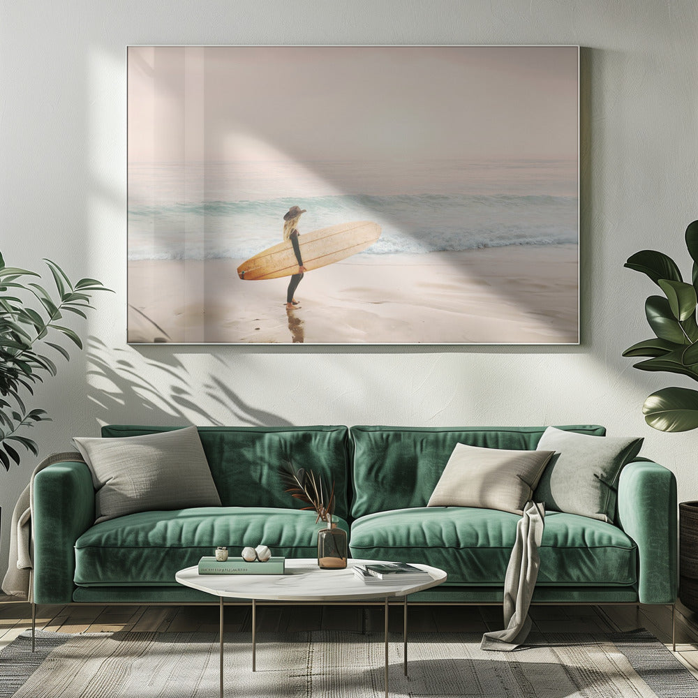 A female surfer with long blonde hair, wearing a black wetsuit and a hat, holds a surfboard while standing on a wet sandy beach looking out at the ocean during a hazy pink sunset. Decor