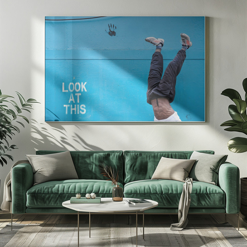 A person's lower body doing a handstand against a bright blue wall with the stenciled words 'LOOK AT THIS' in white. The person wears dark pants and sneakers, and their muscular abdomen is visible. A black handprint is on the wall above the text. The artwork is presented in a silver frame. Print