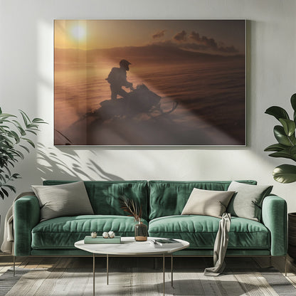 A silhouette of a person wearing a helmet and backpack stands on a snowmobile in a vast, snowy landscape during a golden sunset. The wind blows snow across the ground, creating a hazy, atmospheric effect, with mountains visible in the distance under a warm sky. Wall Art