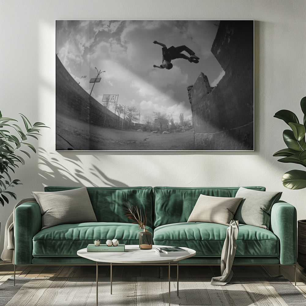 A dynamic black and white fisheye photograph of a person mid-flip, jumping over a crumbling wall in an urban setting under a cloudy sky. The image has a silver-colored frame. Print