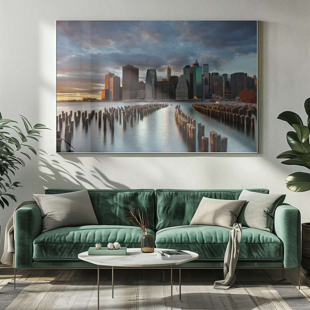 A framed photograph of the Lower Manhattan skyline at sunset, viewed from across the water. Rows of old wooden pier pilings lead the eye towards the city's skyscrapers, which are illuminated by the warm light of the setting sun under a dramatic, cloudy sky. Decor