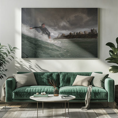 A close-up, low-angle shot of a male surfer in a black wetsuit riding a dark green ocean wave. In the background, a city skyline sits atop a cliff under a dramatic, dark, and stormy sky. The image is presented within a silver picture frame. Print
