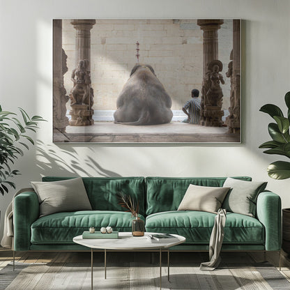 A framed photograph from behind a large elephant sitting on a stone floor inside a temple, flanked by ornate, carved stone pillars. A man in a striped shirt sits on the floor to the right of the elephant, looking towards it. Decor