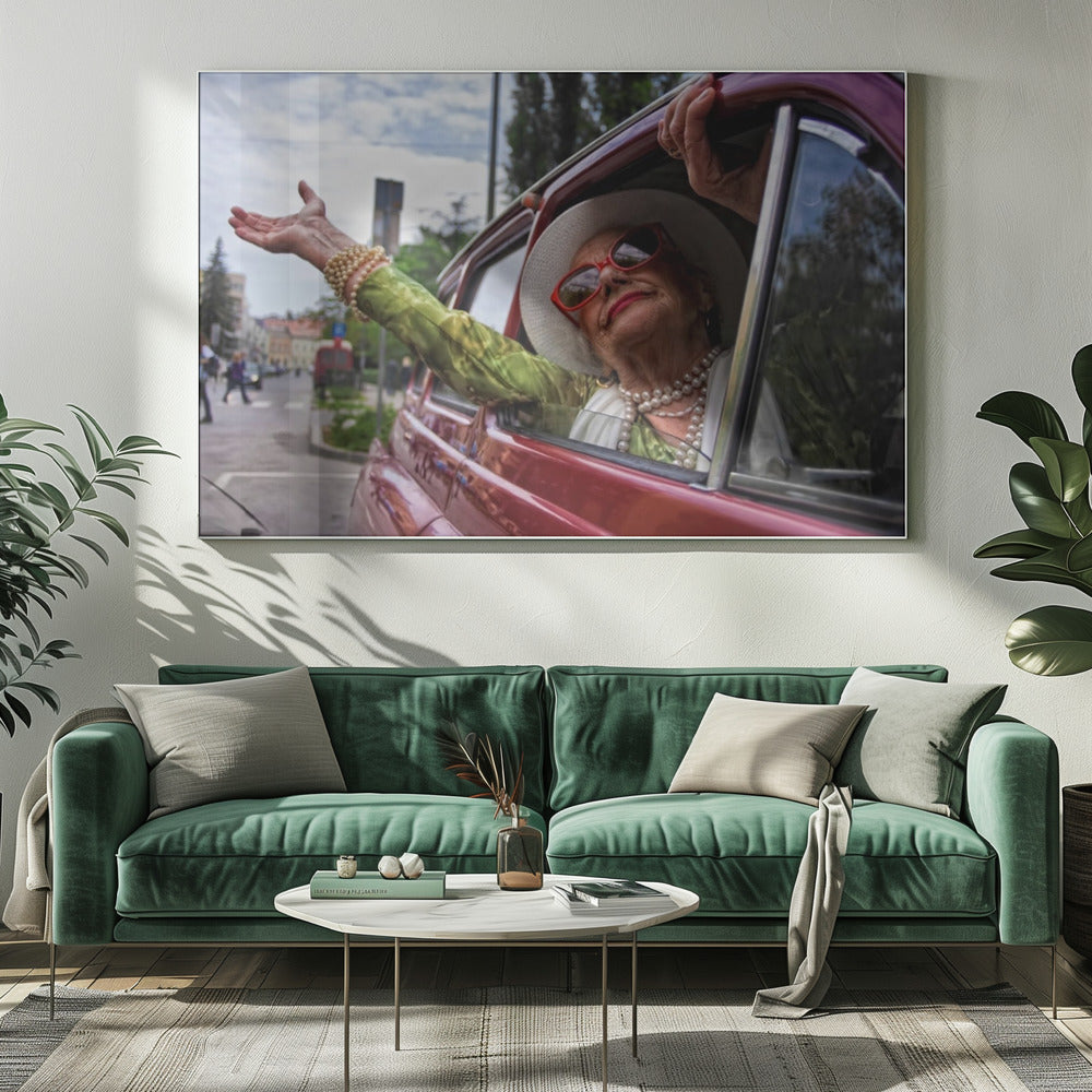A stylish elderly woman wearing a white hat, red sunglasses, and pearl necklaces leans out of the passenger window of a classic red car. Her arm is outstretched with a joyful expression on her face, enjoying a ride through a city street. Poster