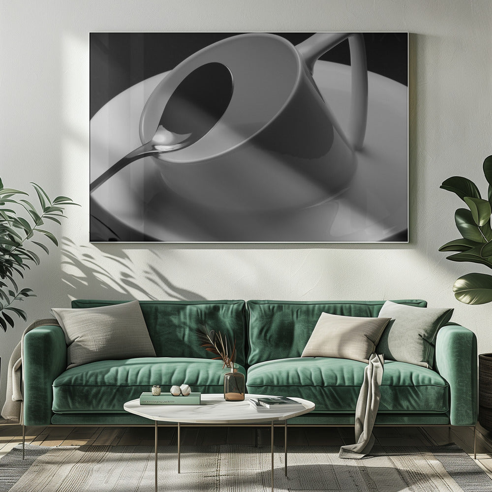 A dramatic black and white close-up photograph of a white porcelain coffee cup tilted on its saucer, with a reflective spoon resting inside. The image has high contrast against a dark background and is presented within a silver-colored frame. Decor