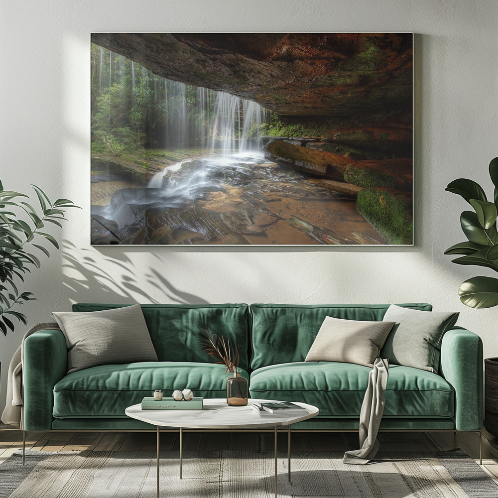 A long-exposure photograph taken from underneath a rock overhang, looking out through a cascading waterfall. The water appears silky as it flows over mossy rocks in a stream below. The scene is framed by a silver border. Print
