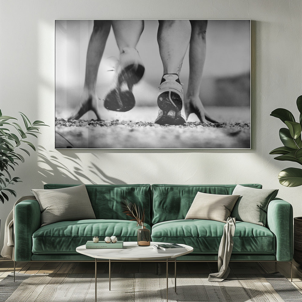 A low-angle, black and white action shot of a runner's legs and hands at the starting position on a gravel track. One foot is planted firmly while the other is in motion, blurred as they push off to start a race. Decor