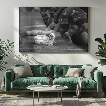 A dynamic black and white, low-angle photograph capturing the intensity of a rugby ruck. A Gilbert rugby ball sits on the turf in the foreground, surrounded by a tangle of players' muscular legs, arms, and cleats, conveying the raw power and action of the sport. Wall Art