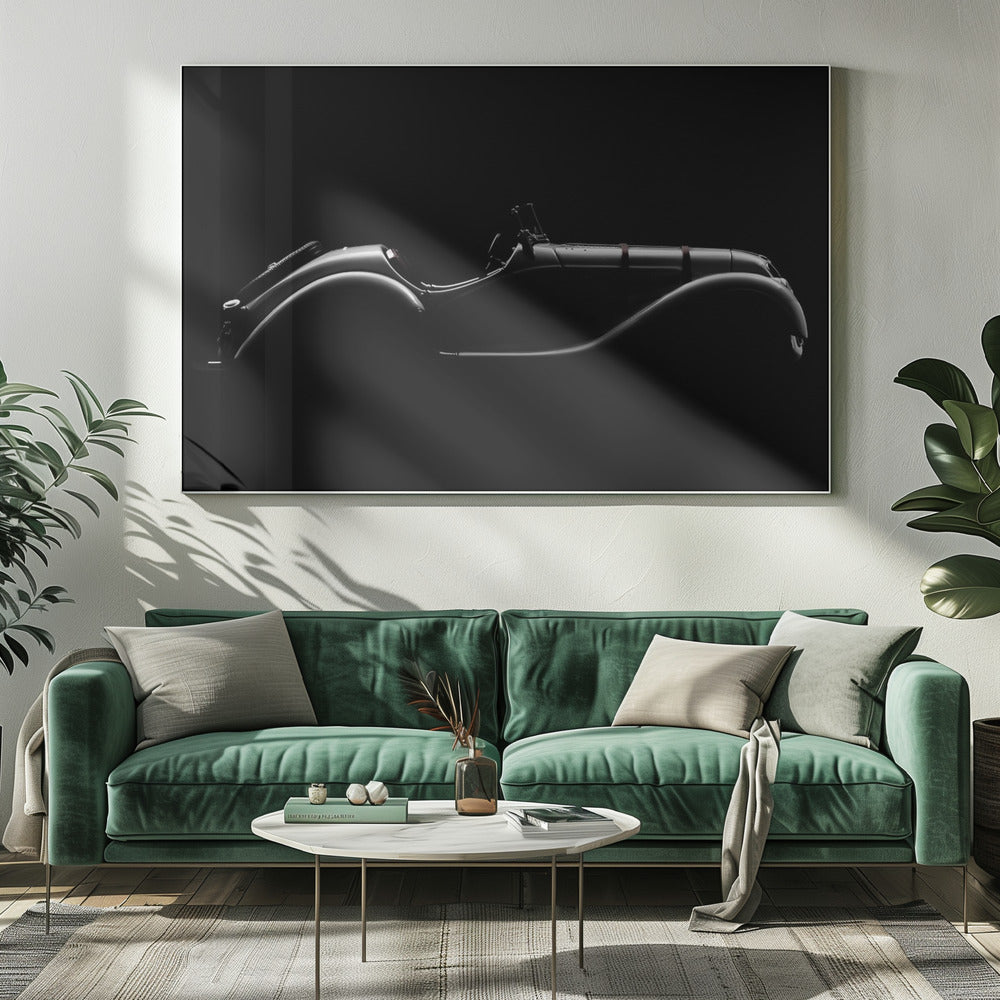 A dramatic black and white photograph showcasing the elegant silhouette of a vintage roadster. The side profile of the classic car is highlighted by subtle light, emphasizing its long hood and sweeping fenders against a solid black background, all within a brushed silver frame. Print
