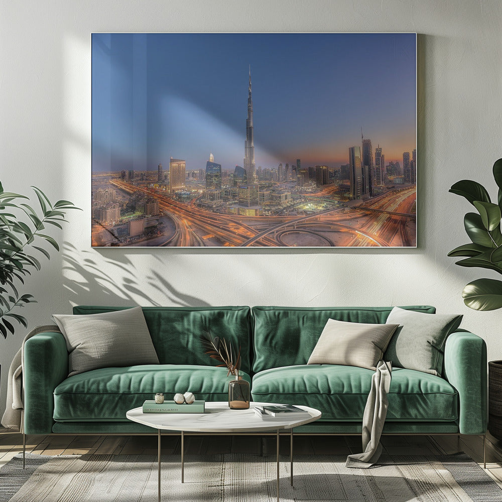 An aerial photograph of the Dubai skyline at dusk, centered on the illuminated Burj Khalifa. The sky transitions from deep blue to warm orange at the horizon, while a complex network of highways below glows with the light trails of traffic. Artwork