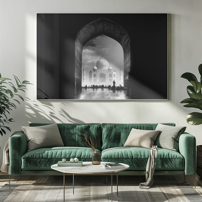 A dramatic black and white photograph of the Taj Mahal seen through a large, dark archway which frames the view. The iconic mausoleum is in the distance under a cloudy sky, and the silhouettes of many people are scattered on the wet, reflective ground in the foreground. Decor