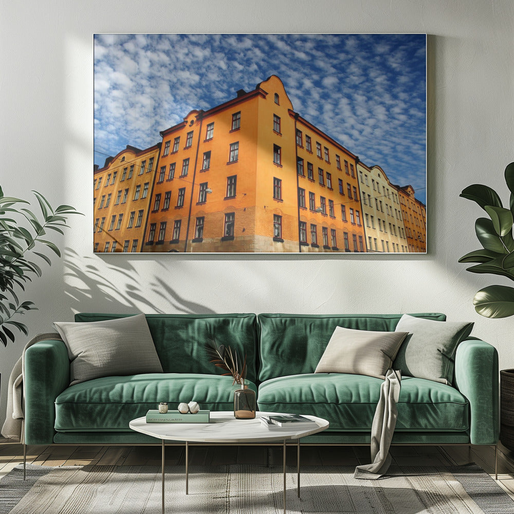 A low-angle shot of a vibrant orange apartment building against a dramatic blue sky filled with small, puffy white clouds. The building is on a corner, with another yellow-hued building visible next to it. The entire image is enclosed in a silver, brushed-metal style frame. Artwork