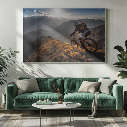 A mountain biker navigates a narrow, golden-lit ridge against a backdrop of a vast mountain range. The sun is breaking through clouds in the distance, casting dramatic rays of light across the epic landscape. Print