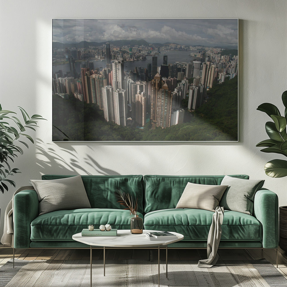 A high-angle photograph of the Hong Kong cityscape, viewed from a lush green peak. Numerous skyscrapers fill the frame, overlooking a bustling harbor with mountains in the distant background under a cloudy sky. The entire image is presented within a silver, brushed-metal frame. Artwork