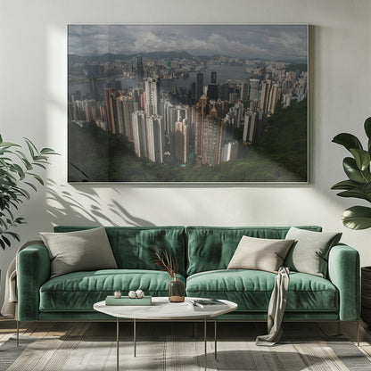 A high-angle photograph of the Hong Kong cityscape, viewed from a lush green peak. Numerous skyscrapers fill the frame, overlooking a bustling harbor with mountains in the distant background under a cloudy sky. The entire image is presented within a silver, brushed-metal frame. Artwork
