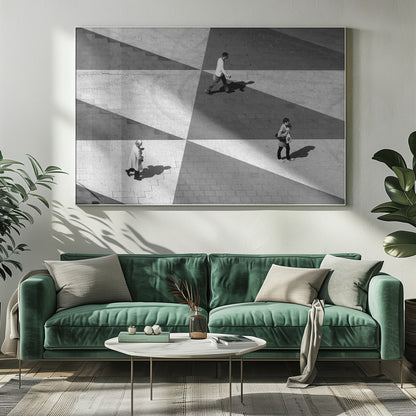 A high-angle, black and white photograph capturing three people walking across a paved plaza, their paths intersecting with a dramatic geometric pattern of light and shadow. The strong sunlight creates large, sharp-edged triangles of shade, isolating each figure in the composition, which is enclosed in a silver frame. Decor