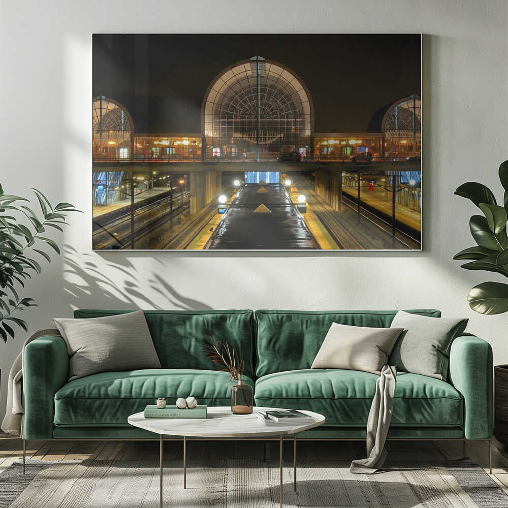 A symmetrical view of a modern train station at night, with a large illuminated arched glass window. Below, empty, wet train tracks and platforms reflect the station's lights, while cars cross an overpass in the middle ground. Wall Art
