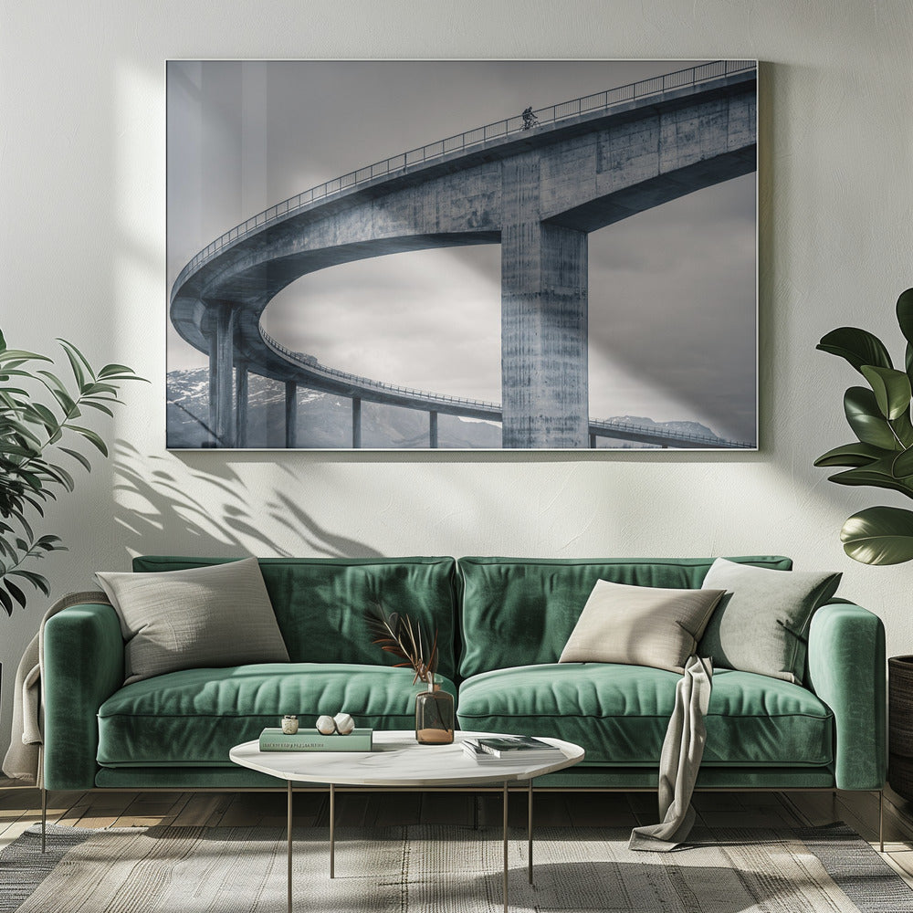A black and white photograph of a lone cyclist riding across the top level of a massive, curving concrete bridge, which sweeps across the frame. In the background, a lower level of the bridge and distant mountains are visible under a cloudy sky. Decor