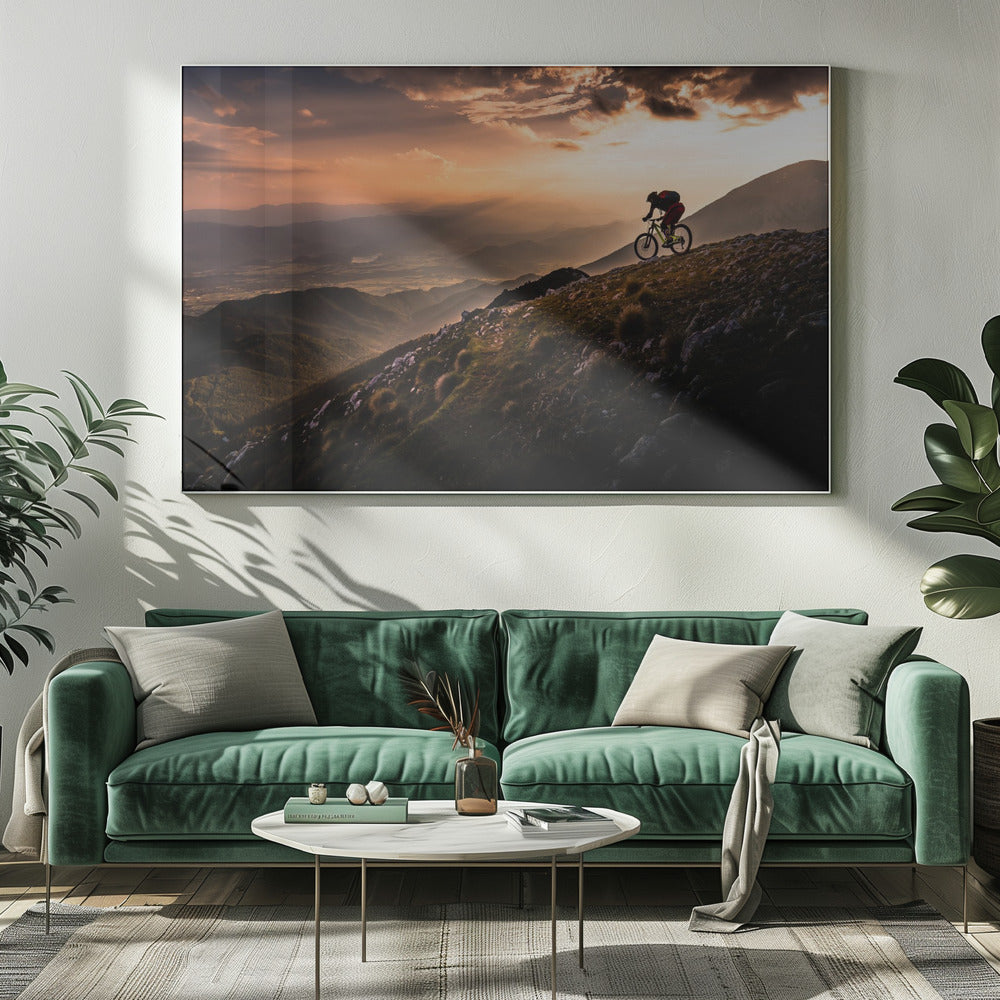 A lone mountain biker is silhouetted against a breathtaking sunset as they navigate a steep, rocky ridge. Below them, layers of mountains and a vast valley stretch into the distance under a dramatic, cloudy orange sky. Artwork