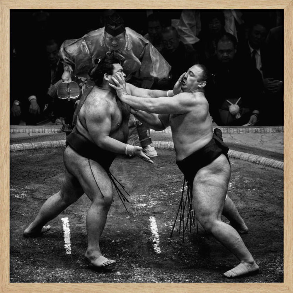 A dynamic black and white action photograph of a sumo wrestling match. Two large wrestlers are in the middle of a clay ring, one pushing forcefully against the other's face. A referee in traditional robes stands behind them, and spectators watch from the background. The entire scene is captured within a light wood frame. Artwork