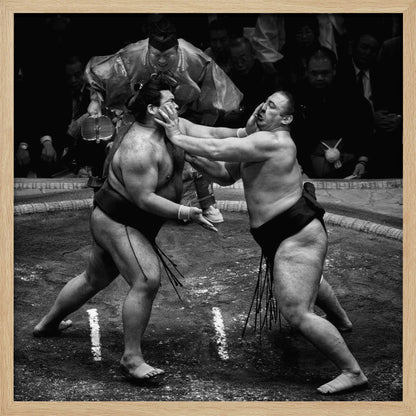 A dynamic black and white action photograph of a sumo wrestling match. Two large wrestlers are in the middle of a clay ring, one pushing forcefully against the other's face. A referee in traditional robes stands behind them, and spectators watch from the background. The entire scene is captured within a light wood frame. Artwork
