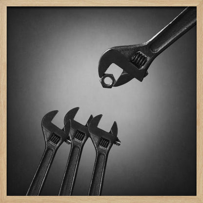 A conceptual black and white photograph in a light wood frame, depicting a large adjustable wrench holding a small nut above three smaller wrenches, which are angled upwards as if they were baby birds waiting to be fed. Print