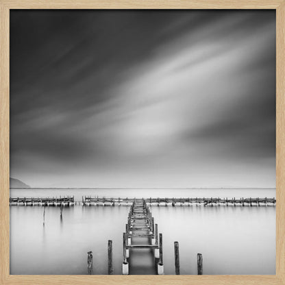 A framed, square black and white long-exposure photograph of a dilapidated wooden pier extending into calm, misty water. The sky is filled with soft, streaked clouds, creating a serene and melancholic mood. Wall Art