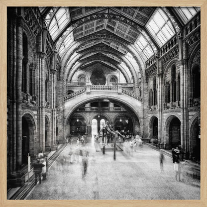 A black and white long exposure photograph of the grand hall of a natural history museum. The ornate, high-arched Romanesque architecture is in sharp focus, while a dinosaur skeleton stands in the center. Crowds of people are blurred into ghostly apparitions as they move through the vast space. Print