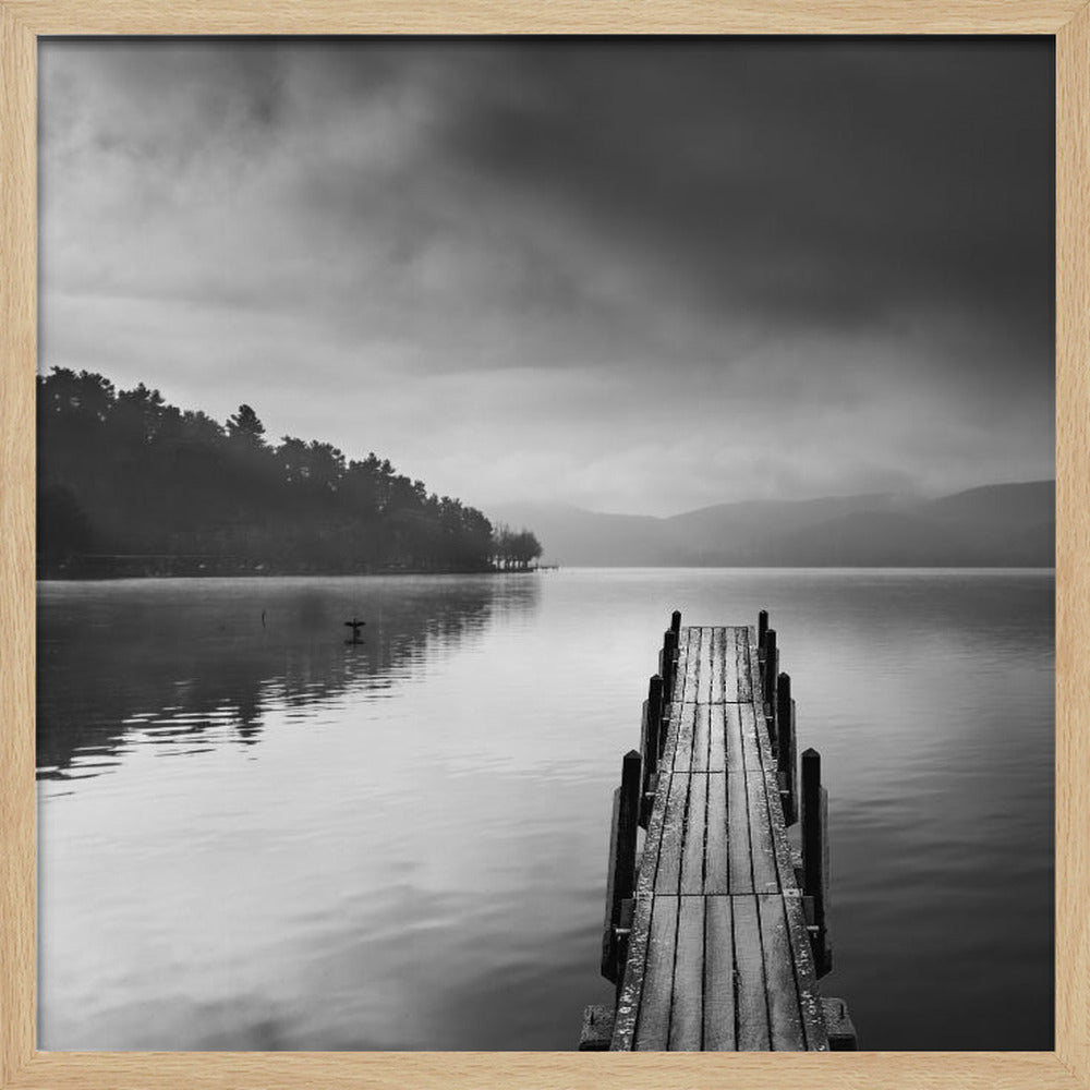 A framed, moody black and white photograph of a wooden pier extending into a calm lake under a cloudy sky, with a forested shoreline and distant mountains in the background. Poster