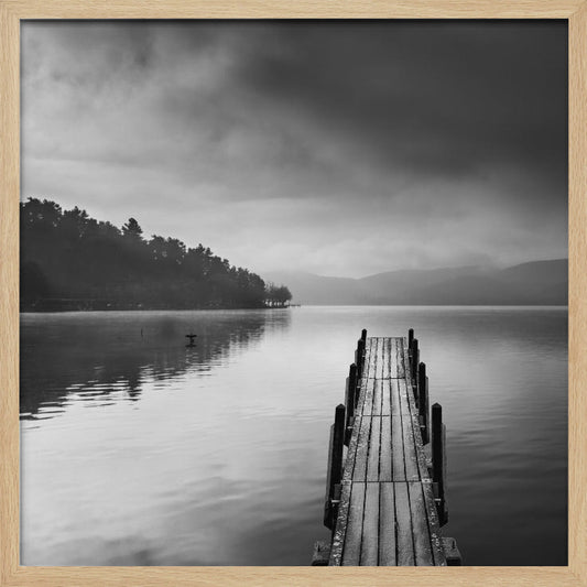 A framed, moody black and white photograph of a wooden pier extending into a calm lake under a cloudy sky, with a forested shoreline and distant mountains in the background. Poster