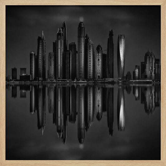 A dramatic black and white photograph of a modern city skyline at night, its towering skyscrapers perfectly reflected in the calm water below, all enclosed within a light brown wooden frame. Print